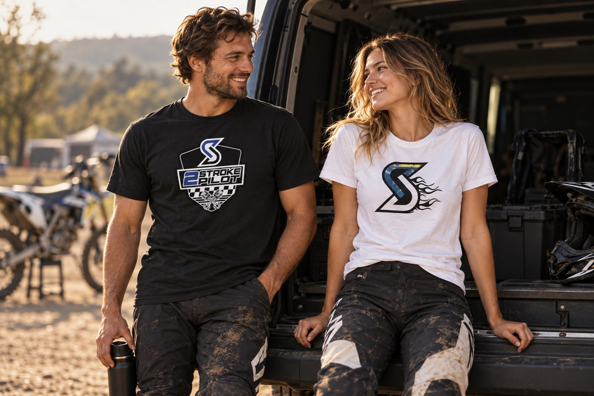 Een man en vrouw dragen het zwarte 2 Stroke Pilot T-shirt en witte Blue Flame T-shirt in een motorcrosspaddock.